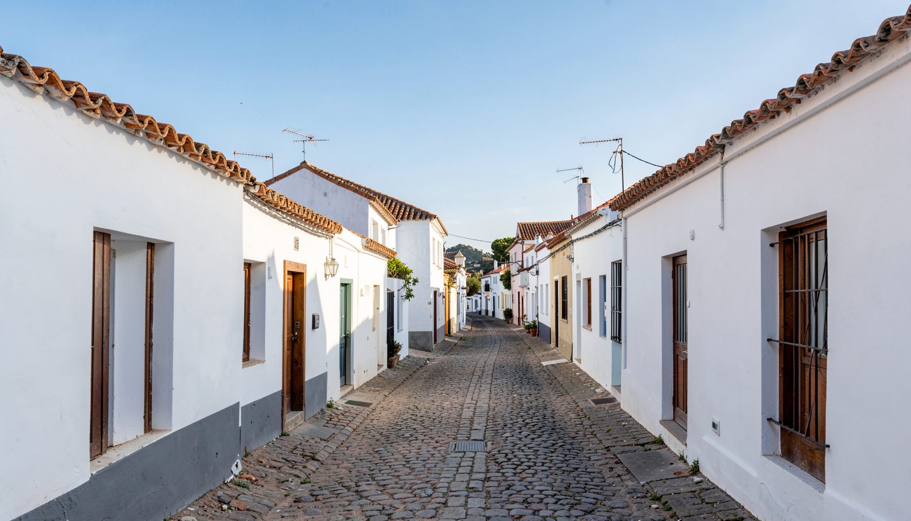 A wide view of a quiet Spanish village street lined with whitewashed houses and terracotta roof tiles, under a bright blue sky