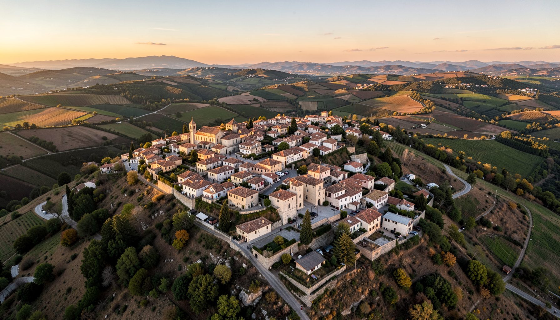 Aerial-style wide view of a hilltop Spanish village at golden hour, with church tower prominent and patchwork fields stretching below
